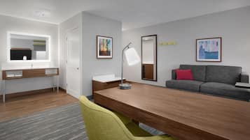 A modern hotel room with a wooden desk, a grey sofa with a red pillow, a large mirror, and two framed artworks on the wall.