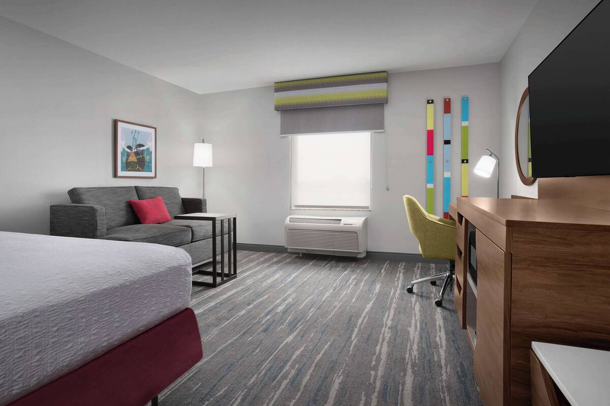 Une chambre d’hôtel comprenant un lit, un canapé, un bureau, une chaise, une télévision, une fenêtre avec des stores, un climatiseur et une lampe.
