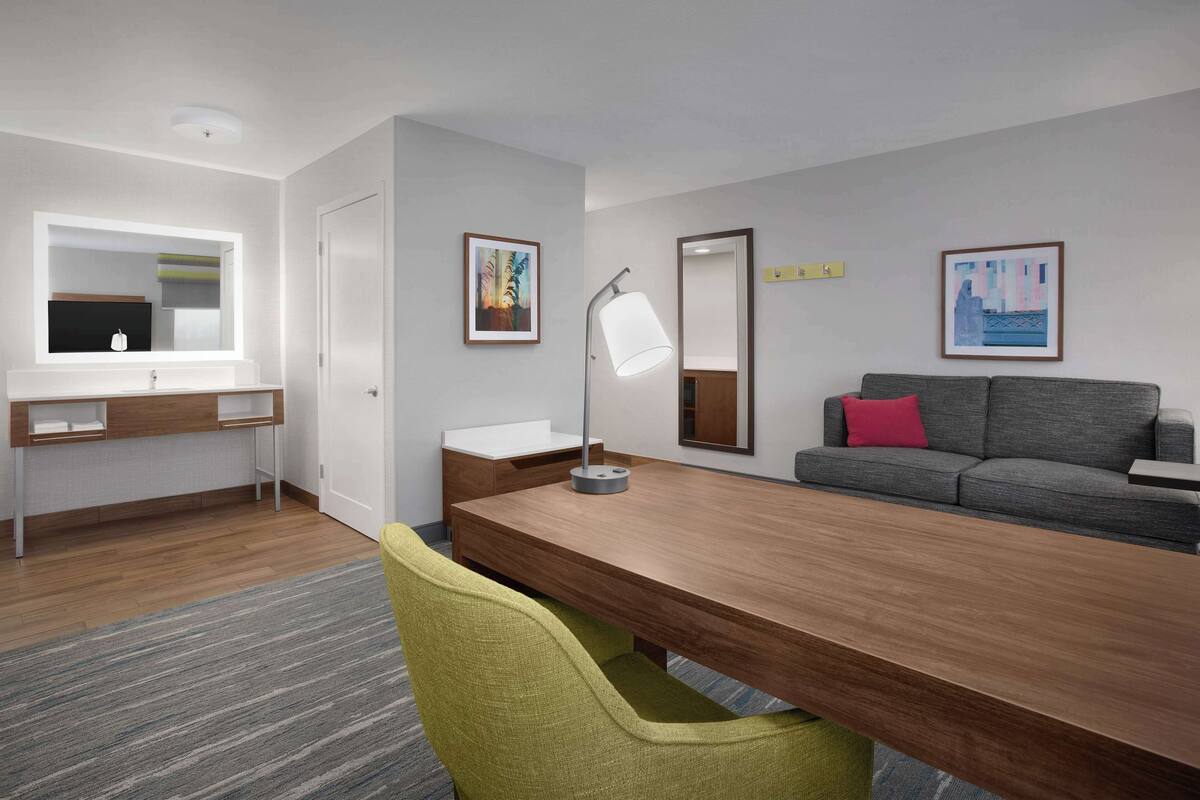 Une chambre d’hôtel moderne avec un bureau en bois, un canapé gris, un miroir et deux œuvres d’art encadrées accrochées au mur.