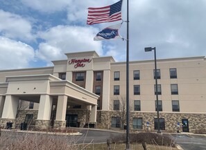 Un hôtel Hampton Inn avec une façade en pierre, une entrée couverte et un drapeau américain.