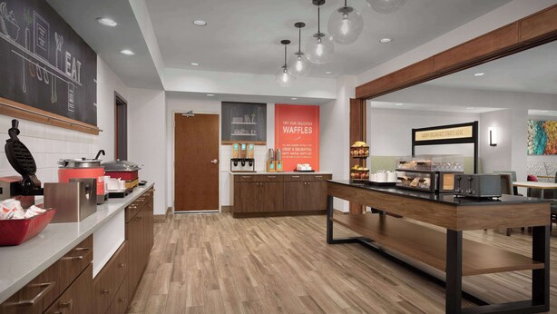 A modern kitchen with wooden flooring, a central island, and a chalkboard with kitchen utensils. There are hanging lights and a sign promoting waffles. - Hampton Inn Yorkville (Yorkville)