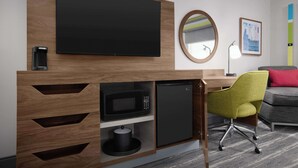 A hotel room with a flat-screen TV mounted above a wooden desk, a microwave, and a mini-fridge. There is a green office chair and a gray sofa with a red pillow. - Hampton Inn Yorkville (Yorkville)