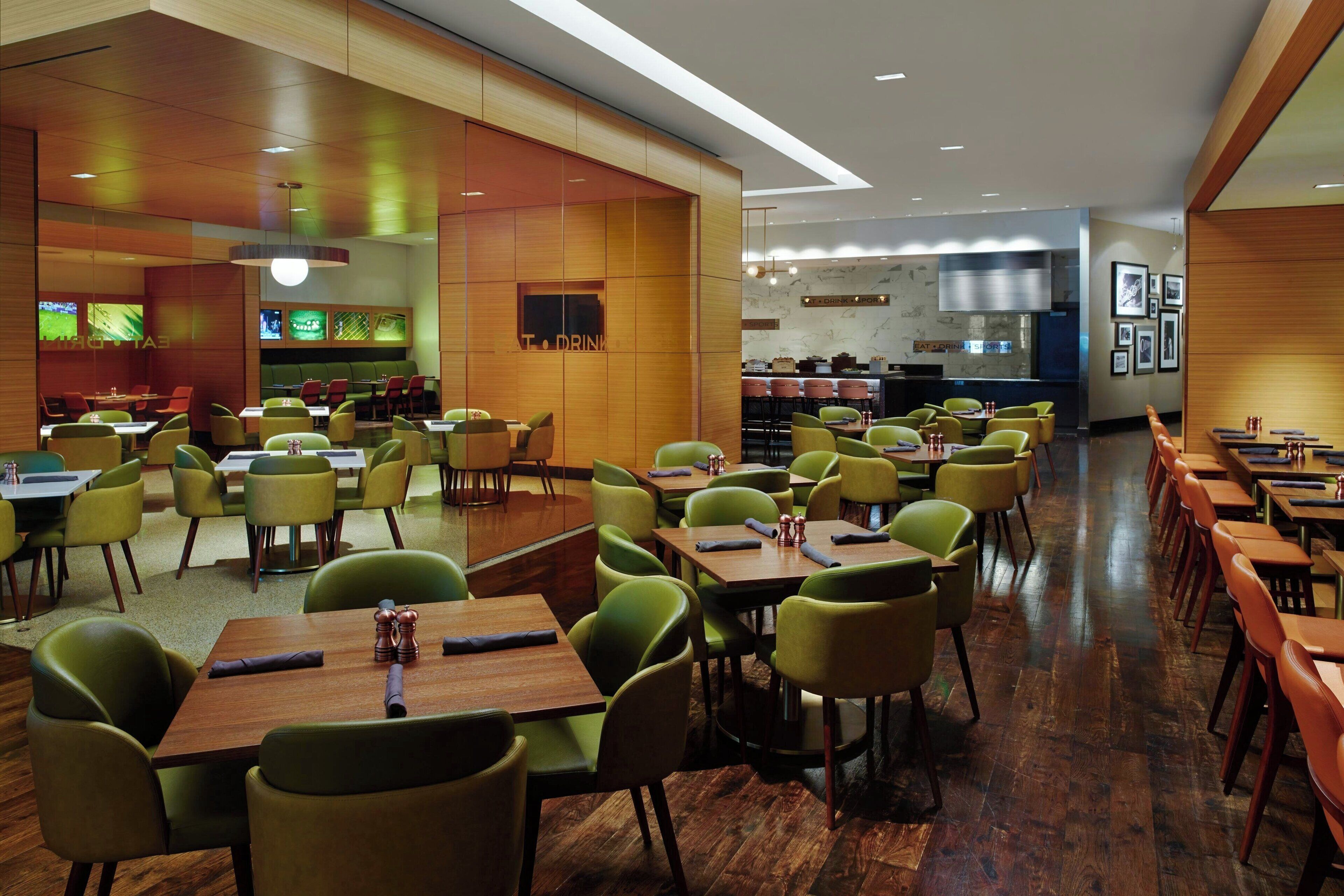 A modern restaurant with green chairs and wooden tables, a bar area, and a kitchen in the background.