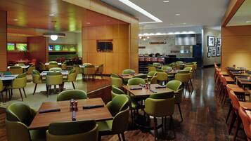 A modern restaurant with green chairs and wooden tables, a bar area, and a kitchen in the background.