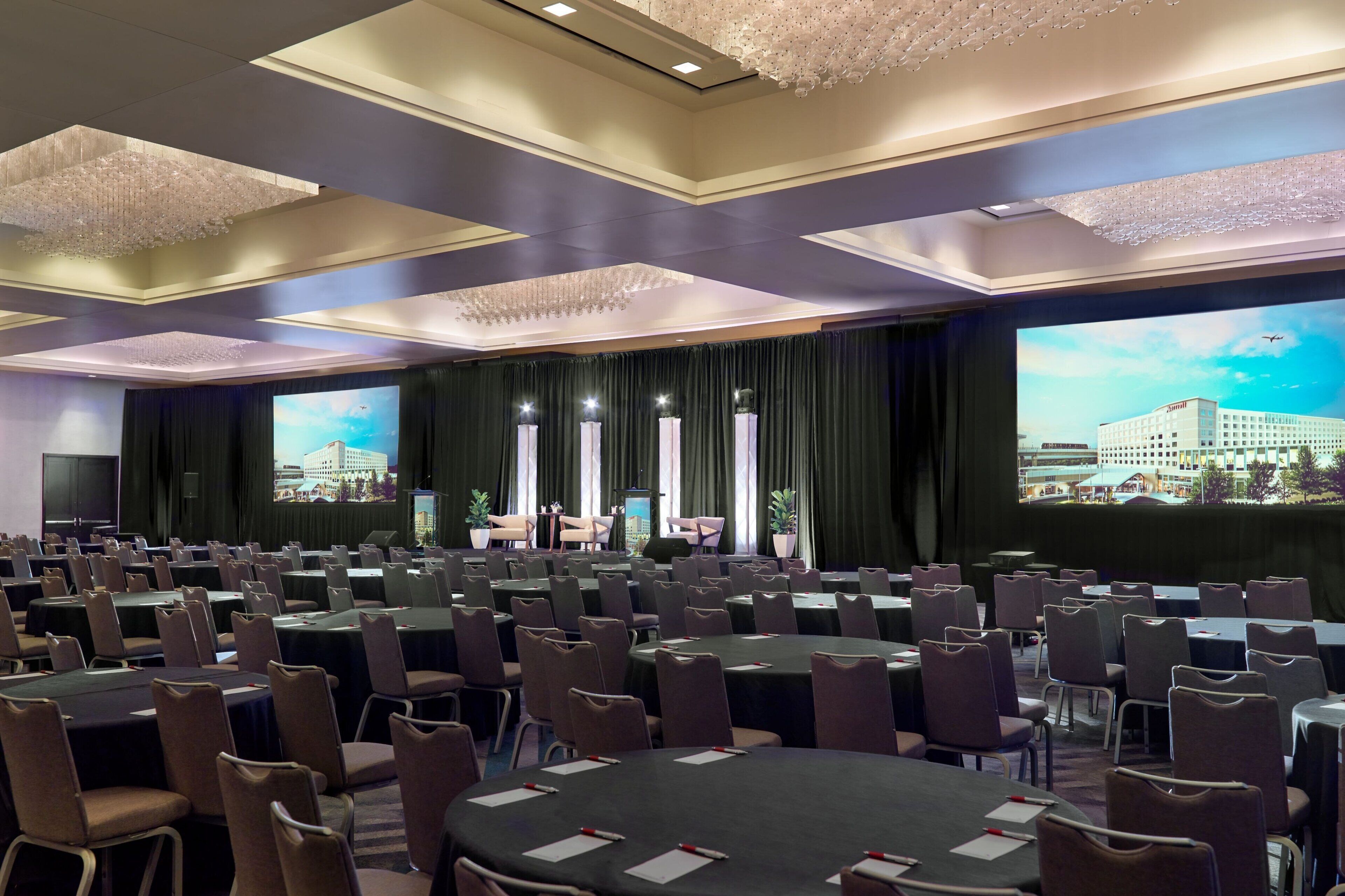 A conference room with multiple tables and chairs, large screens, and a stage with a backdrop.