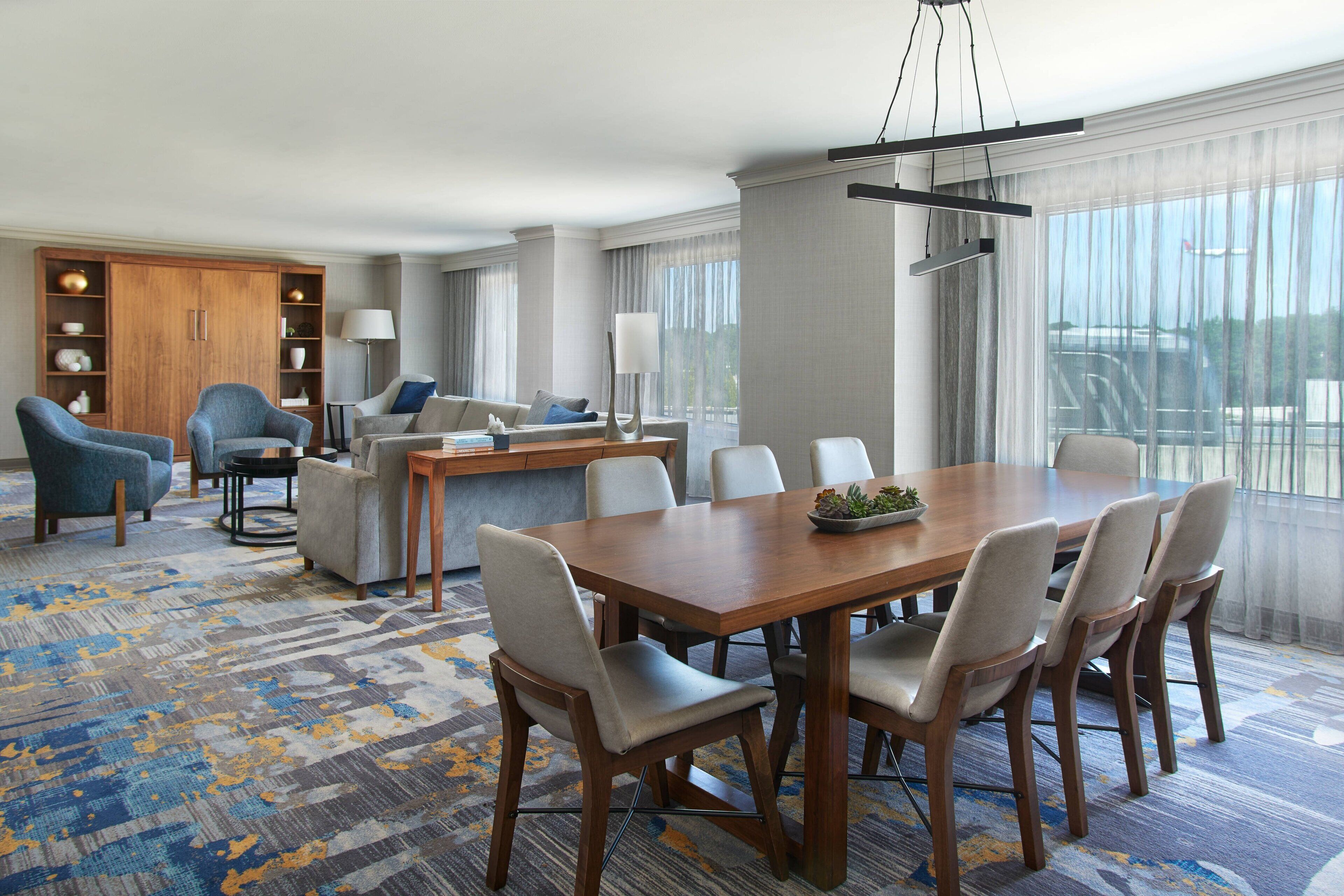 A hotel room with a dining area, a sofa set, a wooden cabinet, and a large window with curtains.