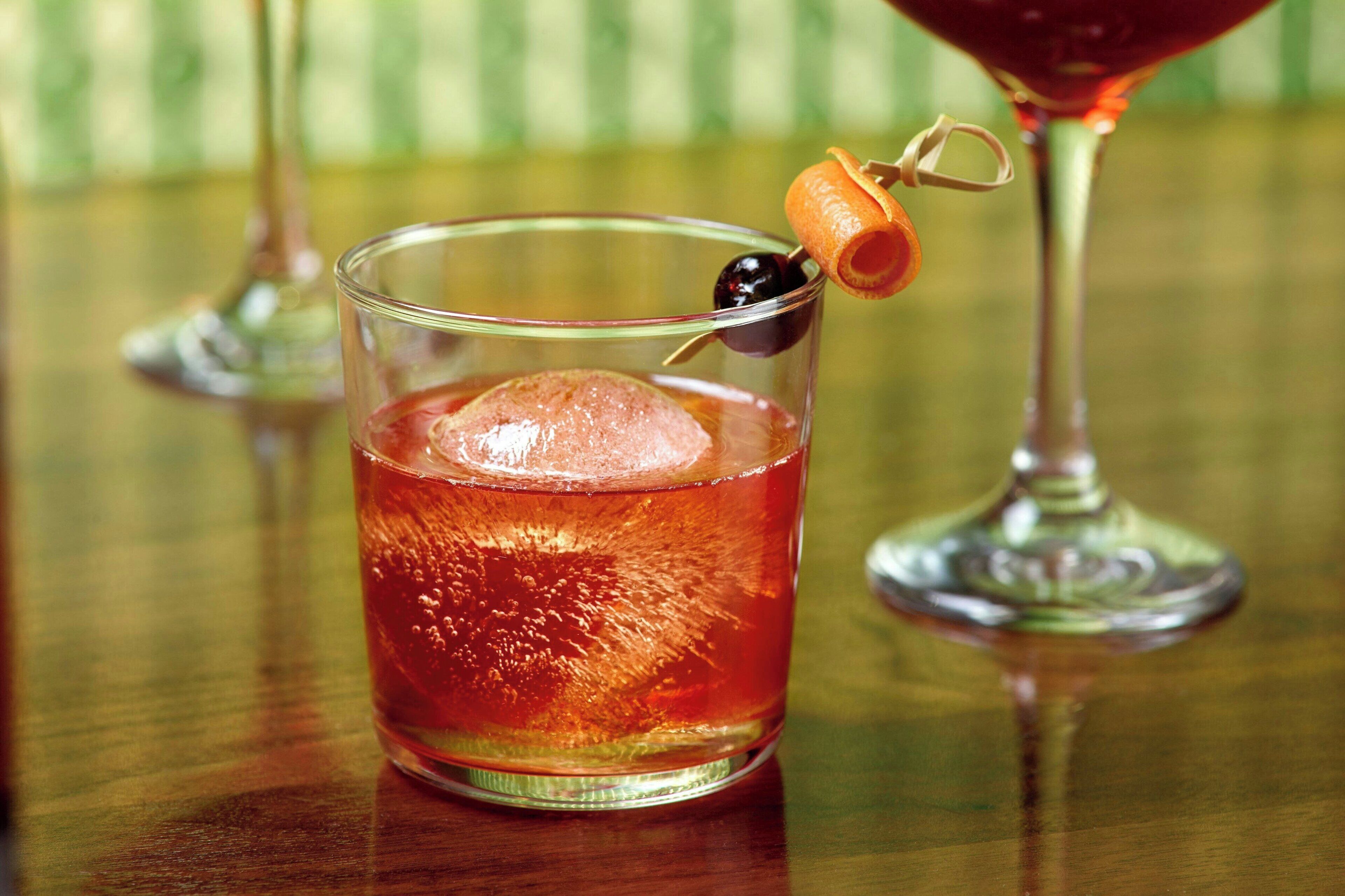 A glass of amber-colored beverage with a cherry and a cherry garnish on a wooden surface.
