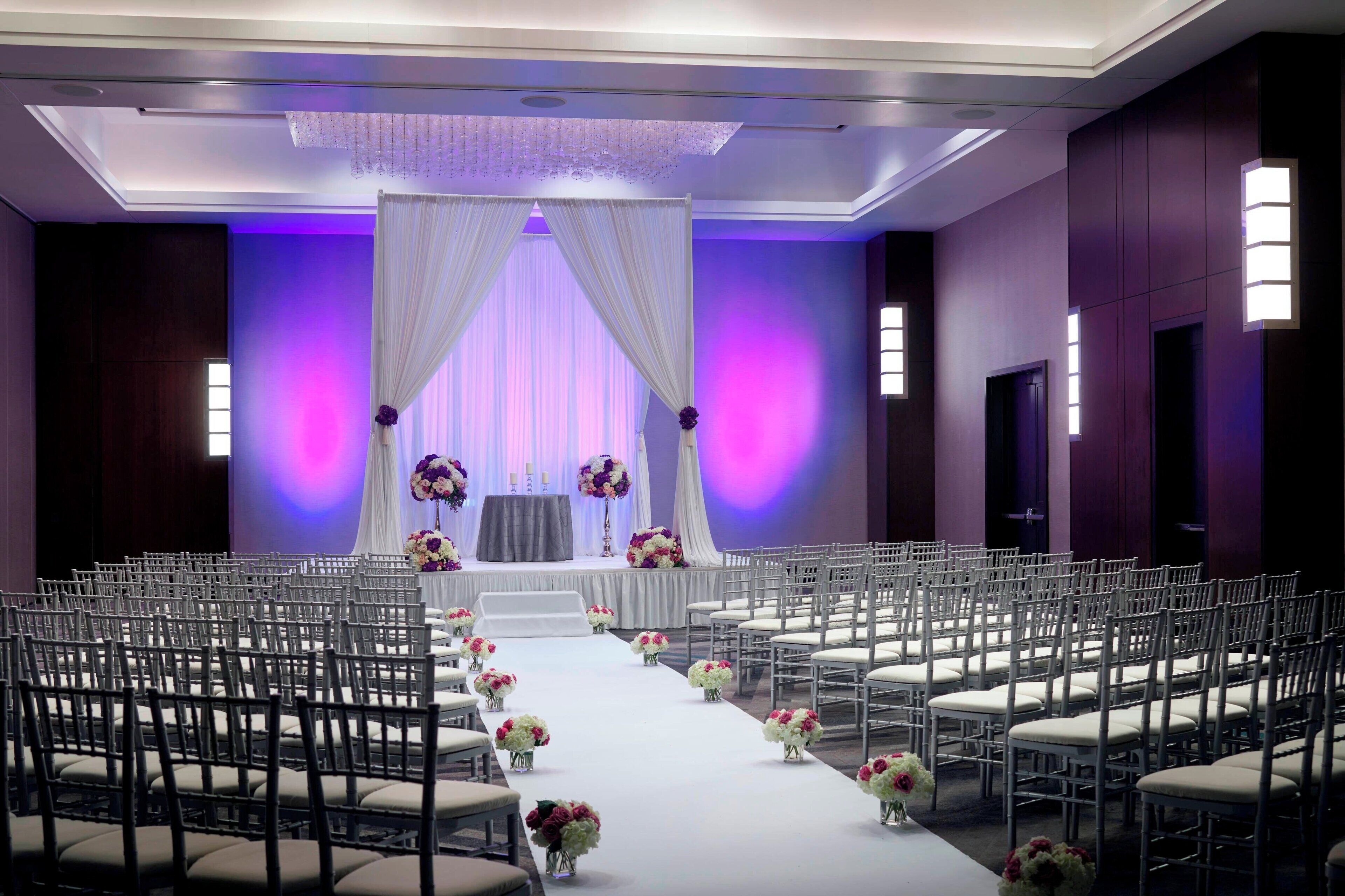 Un espacio para eventos formales con filas de sillas, un escenario con cortinas decorativas y arreglos florales.