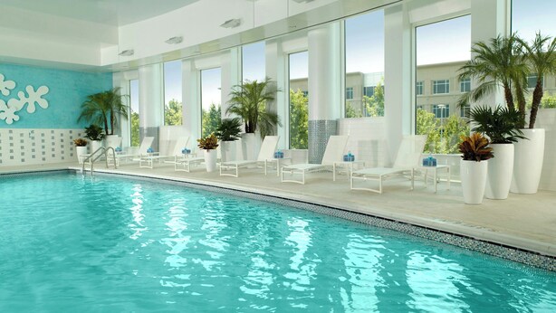A modern indoor swimming pool with clear glass walls, white lounge chairs, and potted plants. - Atlanta Airport Marriott Gateway (College Park)