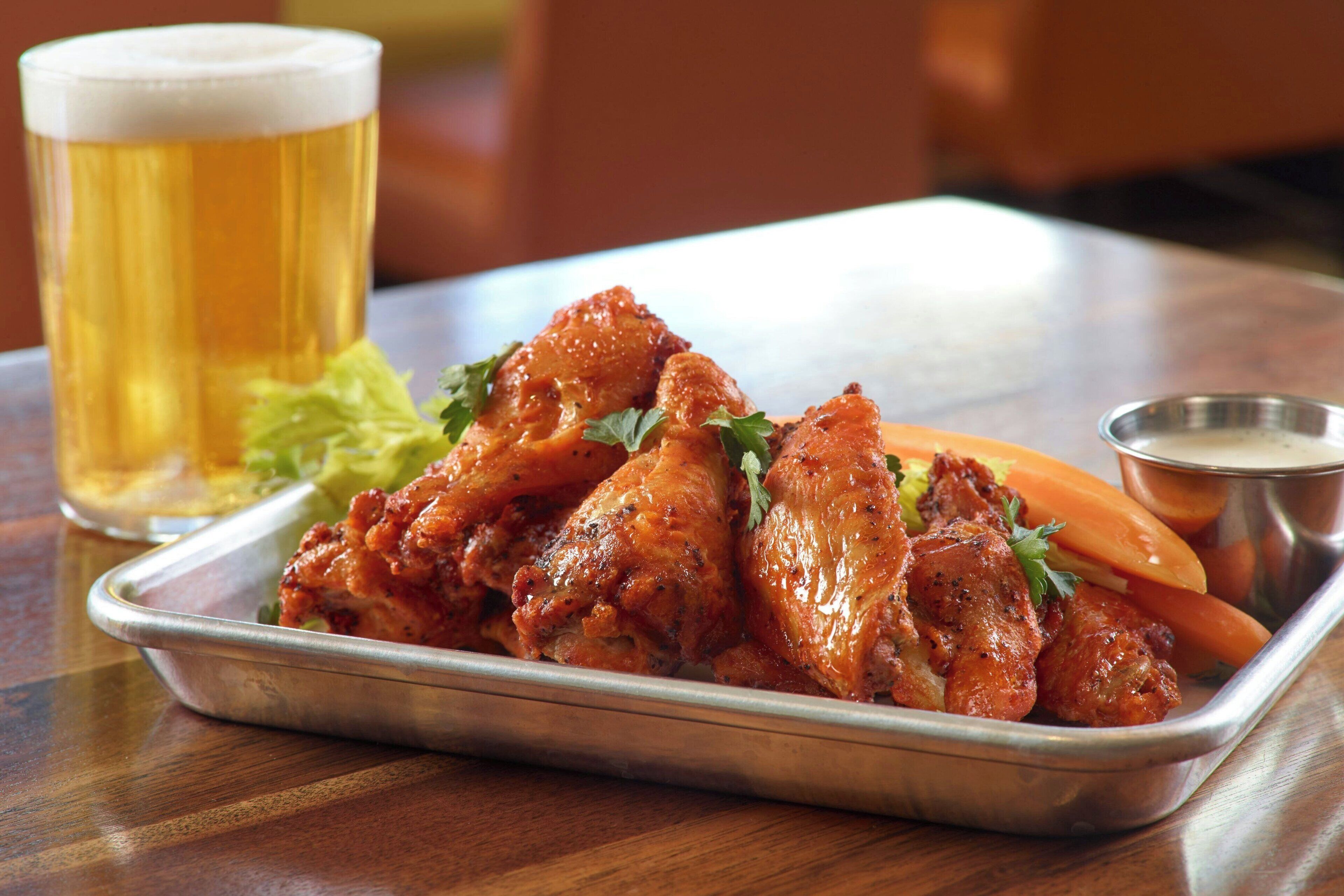 A tray of chicken wings with a side of vegetables and a glass of beer on a wooden table.