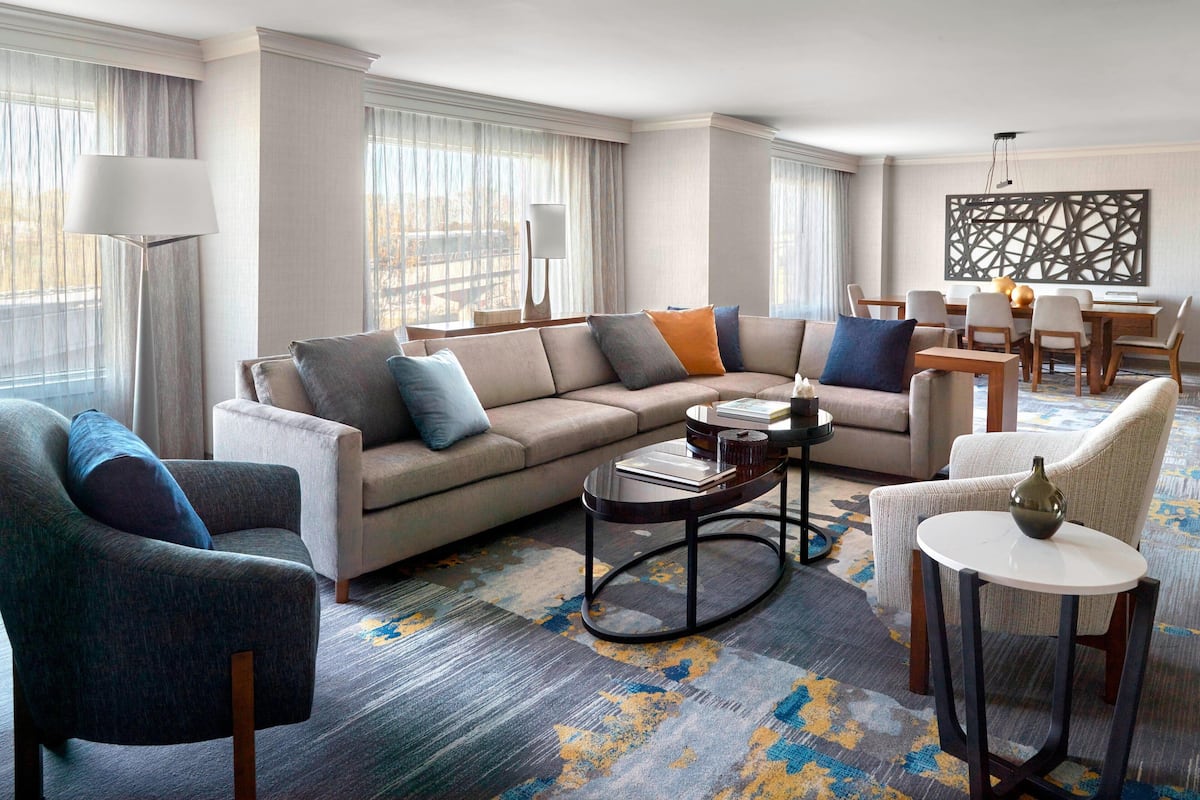 A modern hotel room with a sectional sofa, armchairs, a round coffee table, and a dining area with a chandelier.