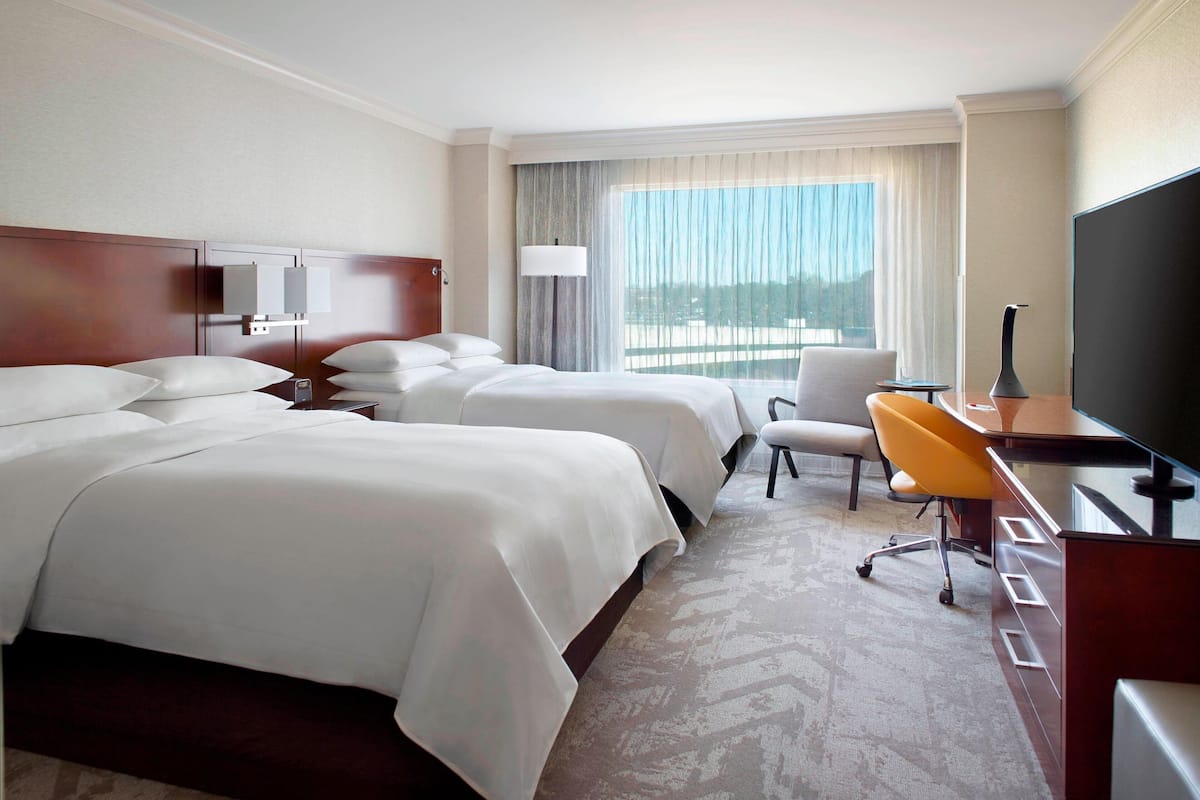 A hotel room with two beds, a desk, a chair, a TV, and a large window with curtains.