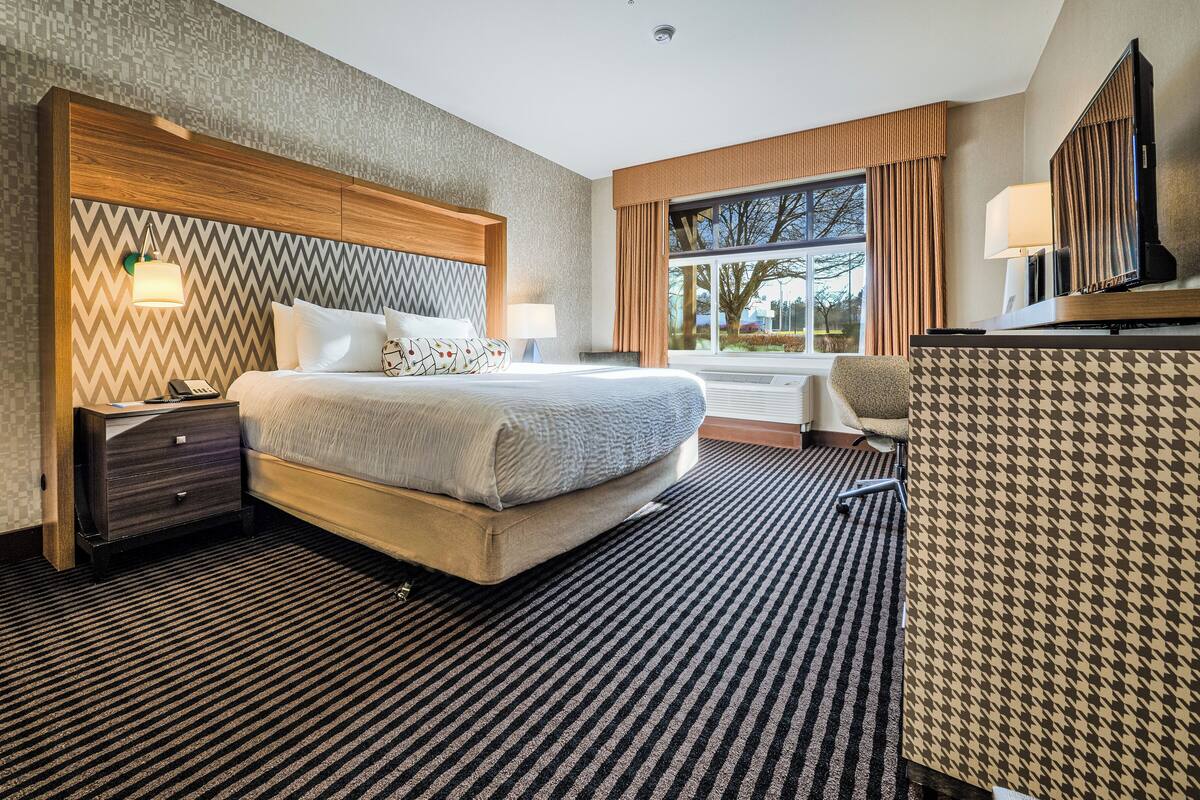A hotel room with a large bed, bedside tables, a desk with a lamp, a chair, and a view of the outdoors.