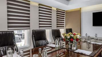 Une salle de conférence avec une table en verre, des fauteuils en cuir, un arrangement floral au centre et un téléviseur à écran plat fixé au mur.