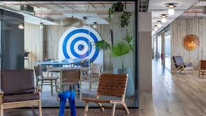 Un moderno spazio ufficio con un'ampia parete vetrata, una decorazione a bersaglio in bianco e blu, e diverse sedie e tavoli di legno.