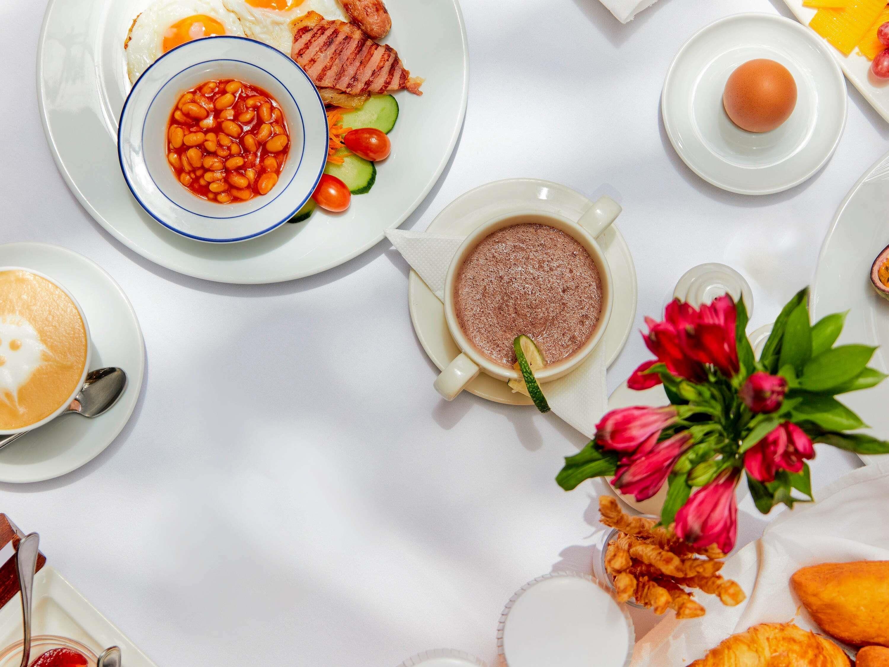 Ein Frühstückstisch mit Eiern, Speck, Bohnen und Gemüse, einer Tasse Kaffee und einer heißen Schokolade mit Zitronenscheibe.