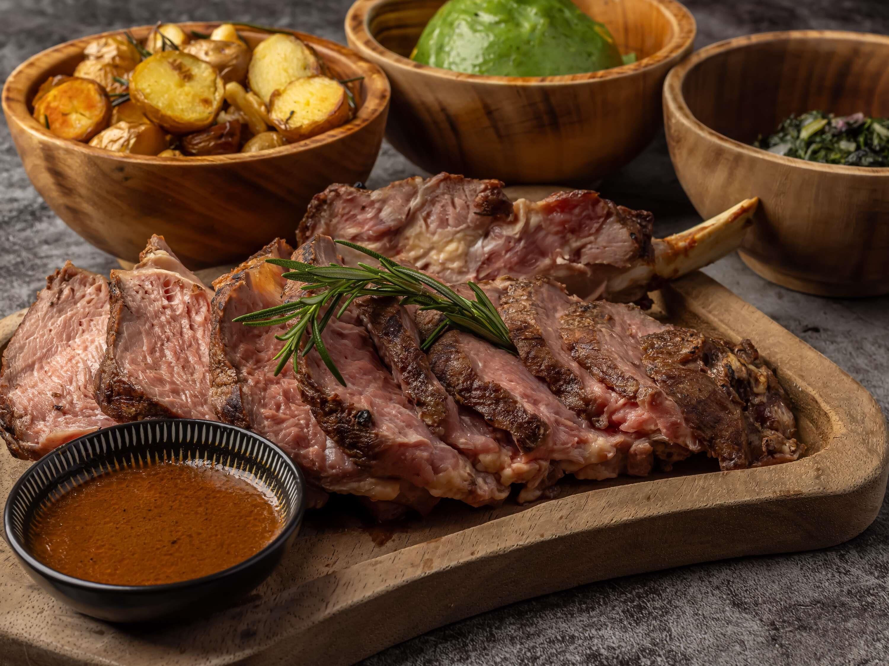 A wooden cutting board with sliced meat, a bowl of roasted potatoes, and a small bowl of sauce.