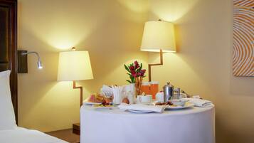 A hotel room with a bed, a round table with breakfast items, two wall-mounted lamps, and a painting on the wall.
