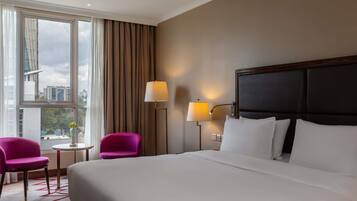 A hotel room with a large bed, two chairs, a small table, and a view of buildings through the window.
