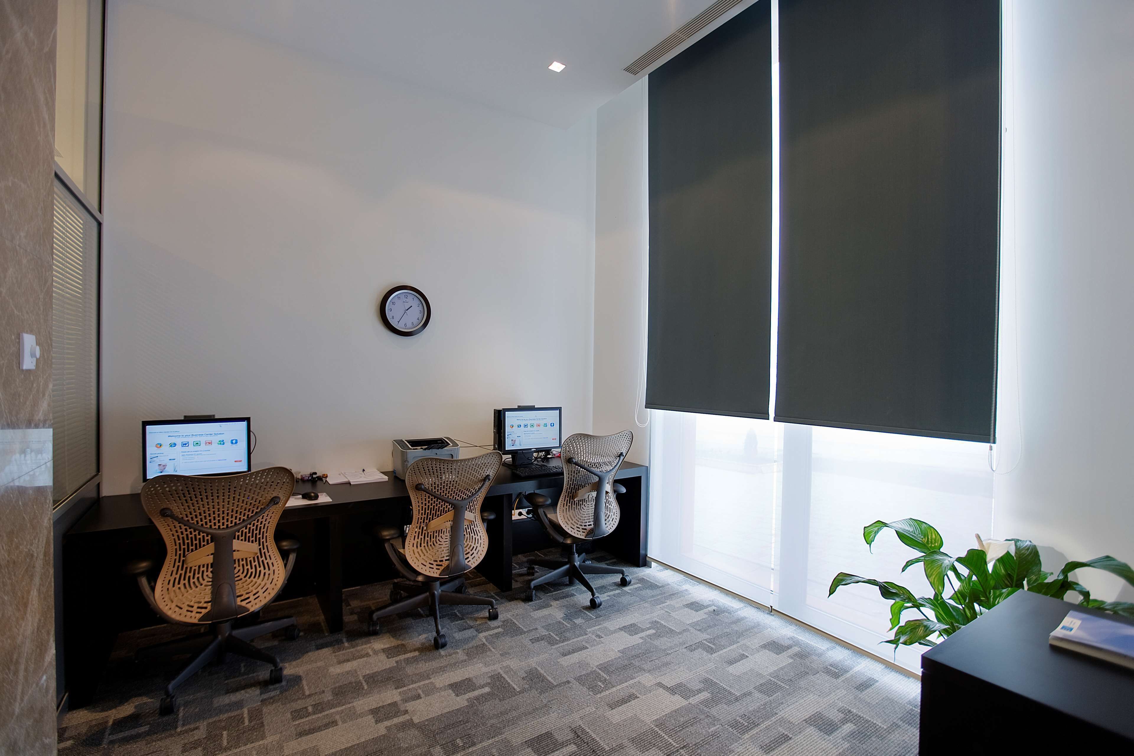 A modern office space with two desks, two office chairs, a clock on the wall, and a large window with blinds.