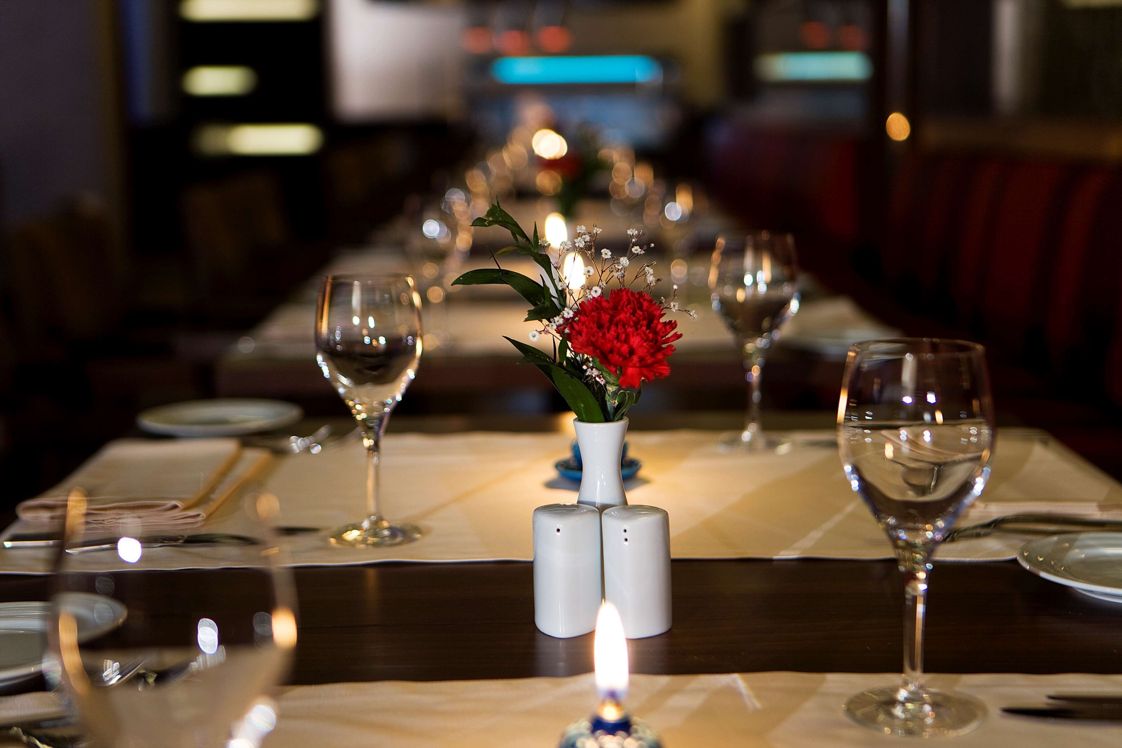 Una mesa de restaurante dispuesta con copas de vino, una vela y un centro de mesa con flores rojas y blancas.