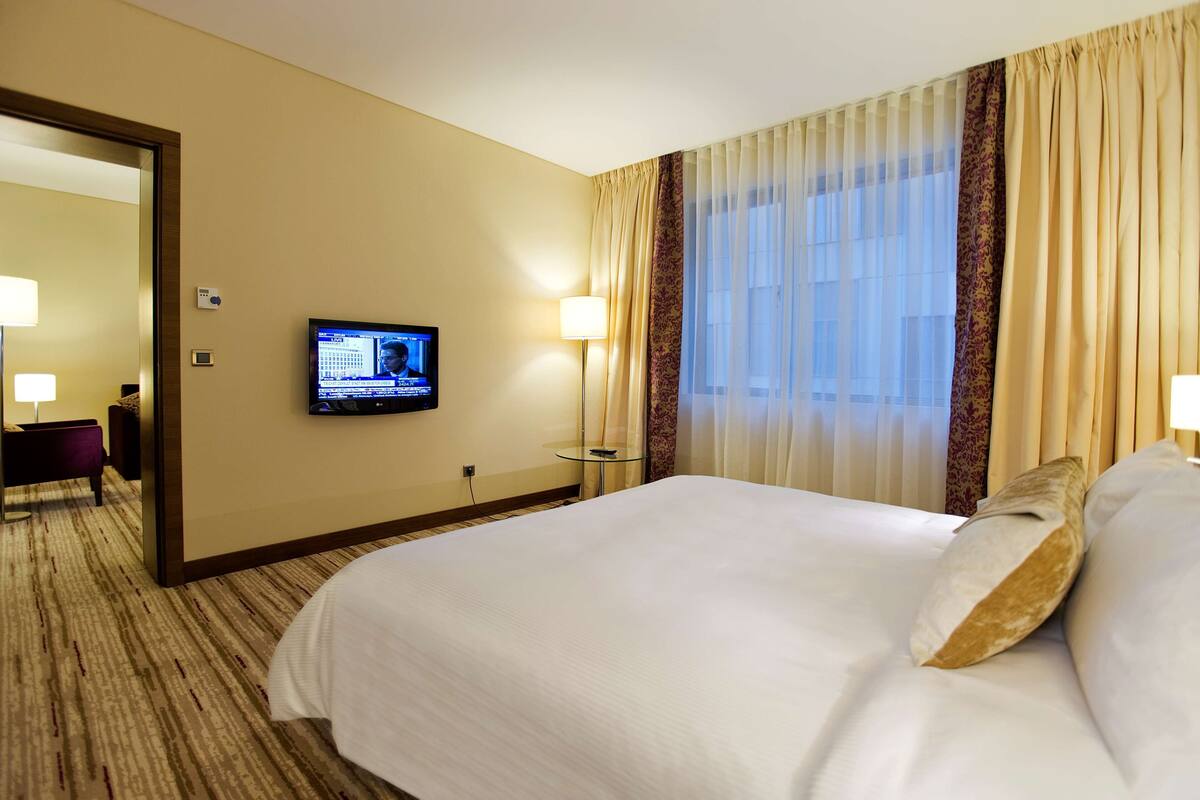 A hotel room with a large bed, a television on the wall, a desk with a chair, and a window with curtains.