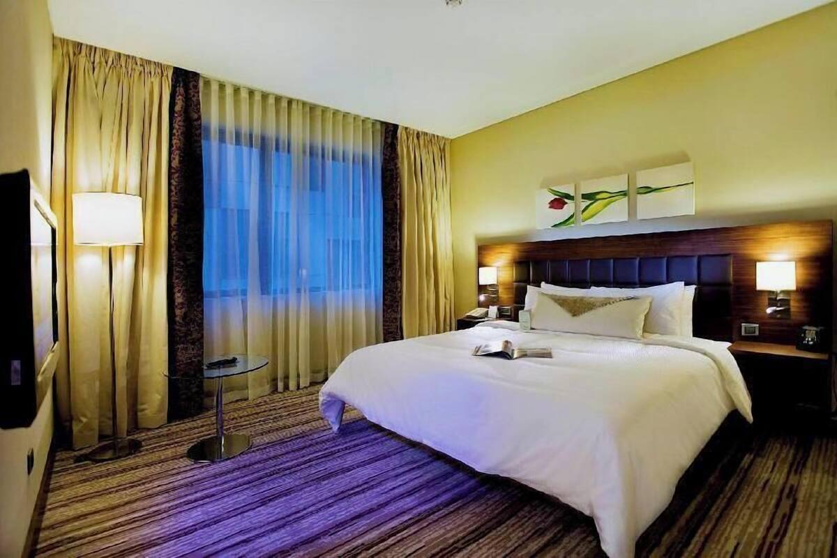 A hotel room with a large bed, a bedside table, a lamp, a window with curtains, and a wall-mounted artwork.