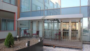 A modern hotel with a glass facade, outdoor seating area, and a small garden.