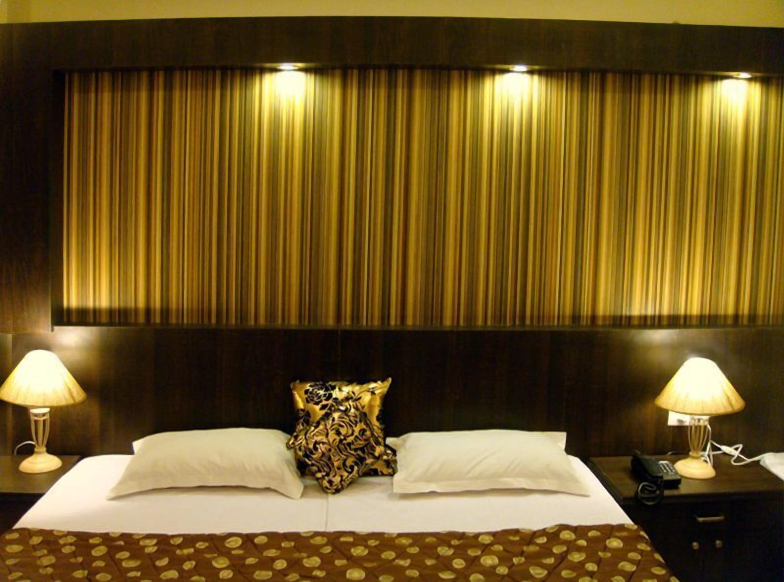 A hotel room with a large bed, two bedside lamps, a patterned pillow, and a dark headboard.