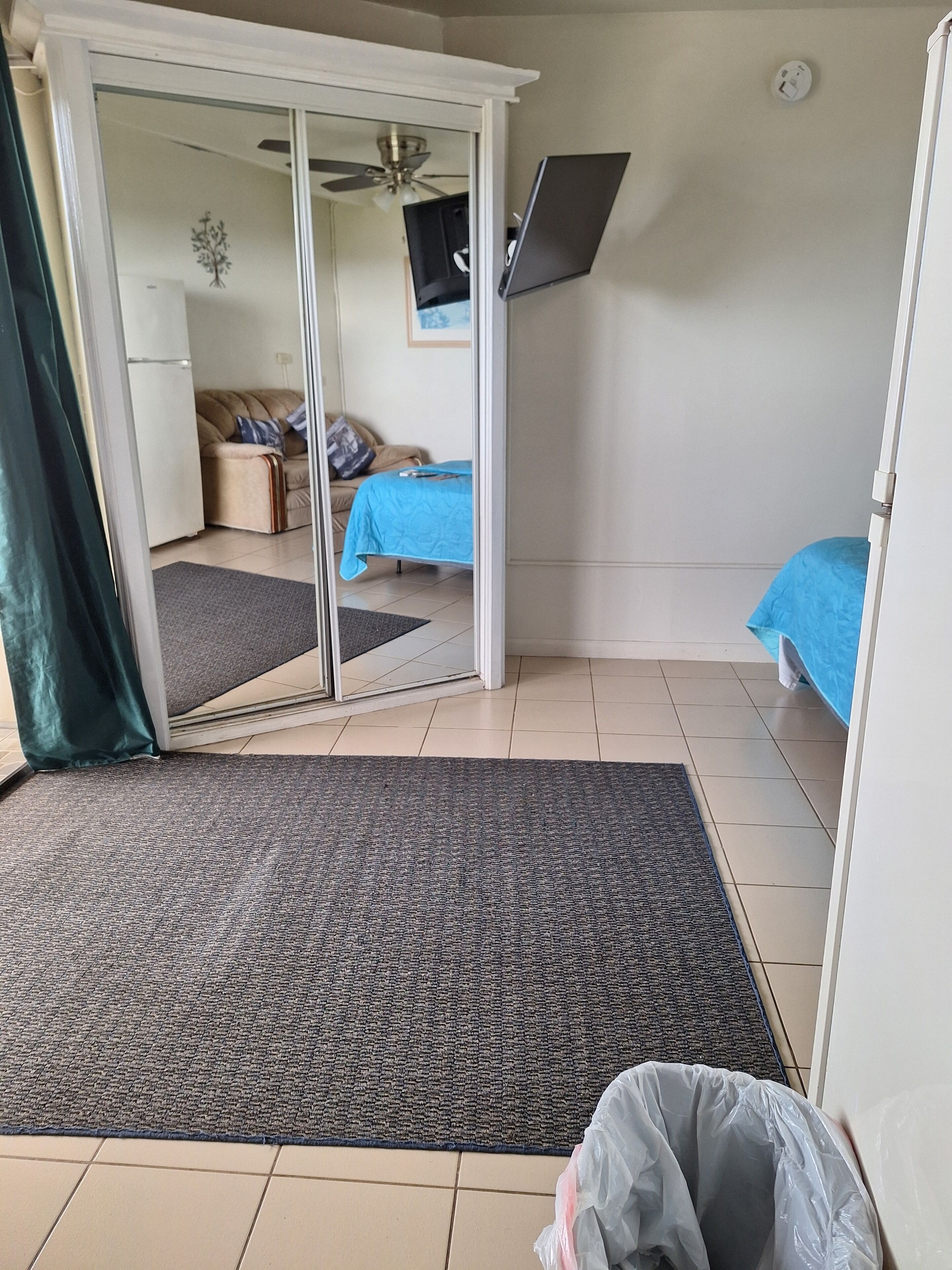 A room with a sliding glass door leading to a living area, a bed with a blue cover, a ceiling fan, and a mounted TV.