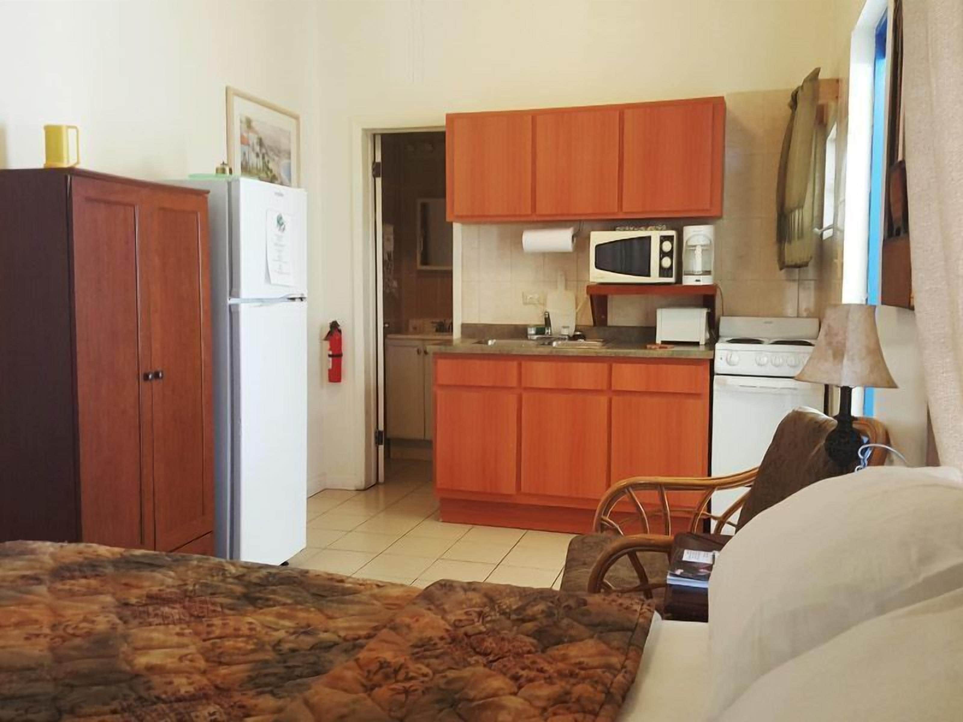 A compact hotel room with a bed, a small kitchenette, a wooden wardrobe, and a door leading to another room.