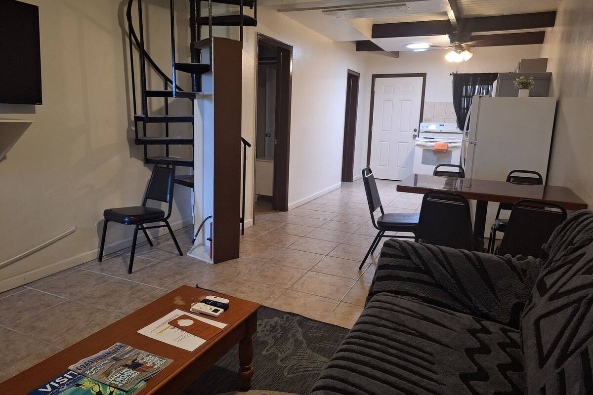 A living room with a sofa, coffee table, and a spiral staircase. There is a dining area with a table and chairs. The room has tiled flooring and a ceiling fan.