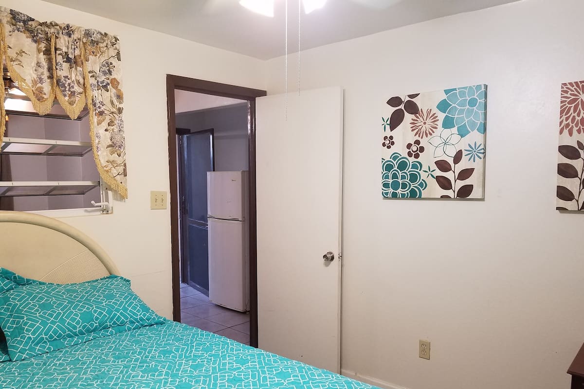 A bedroom with a bed, a door, a refrigerator, and a wall with floral artwork.