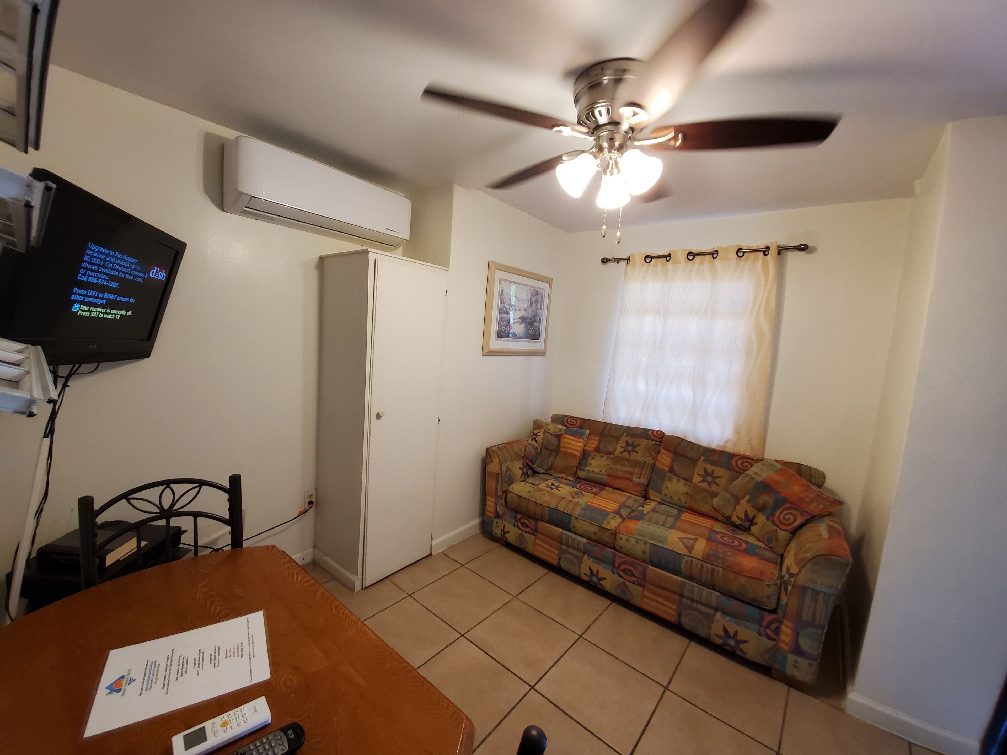 A living room with a ceiling fan, a patterned sofa, a television mounted on the wall, and a small table with a remote control.