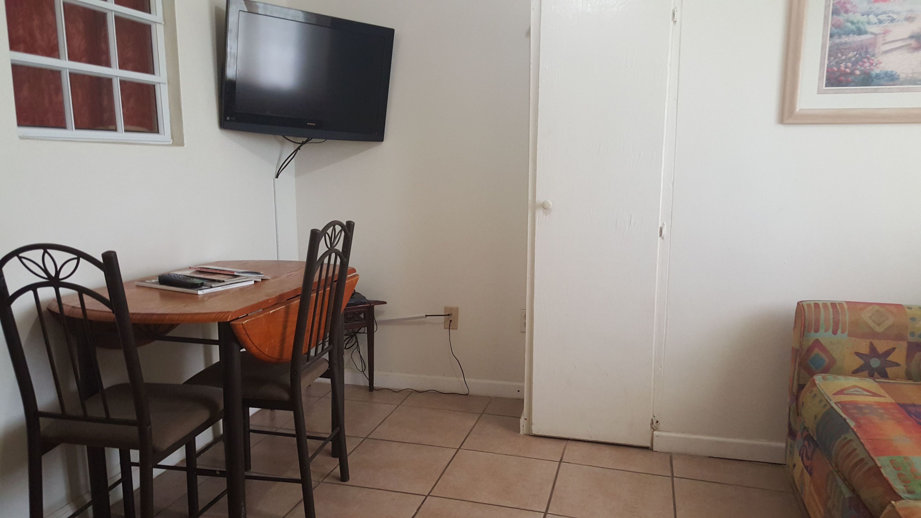 Una habitación pequeña con una mesa de madera, dos sillas, un televisor en la pared y un sofá colorido.