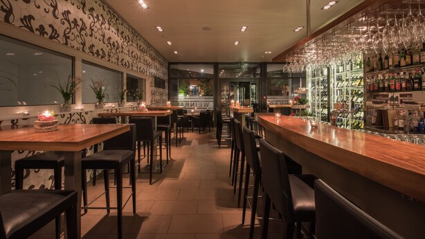A bar area with wooden tables and chairs, a glass display case with bottles, and a patterned wall. - Hotel Arcade (Sins)