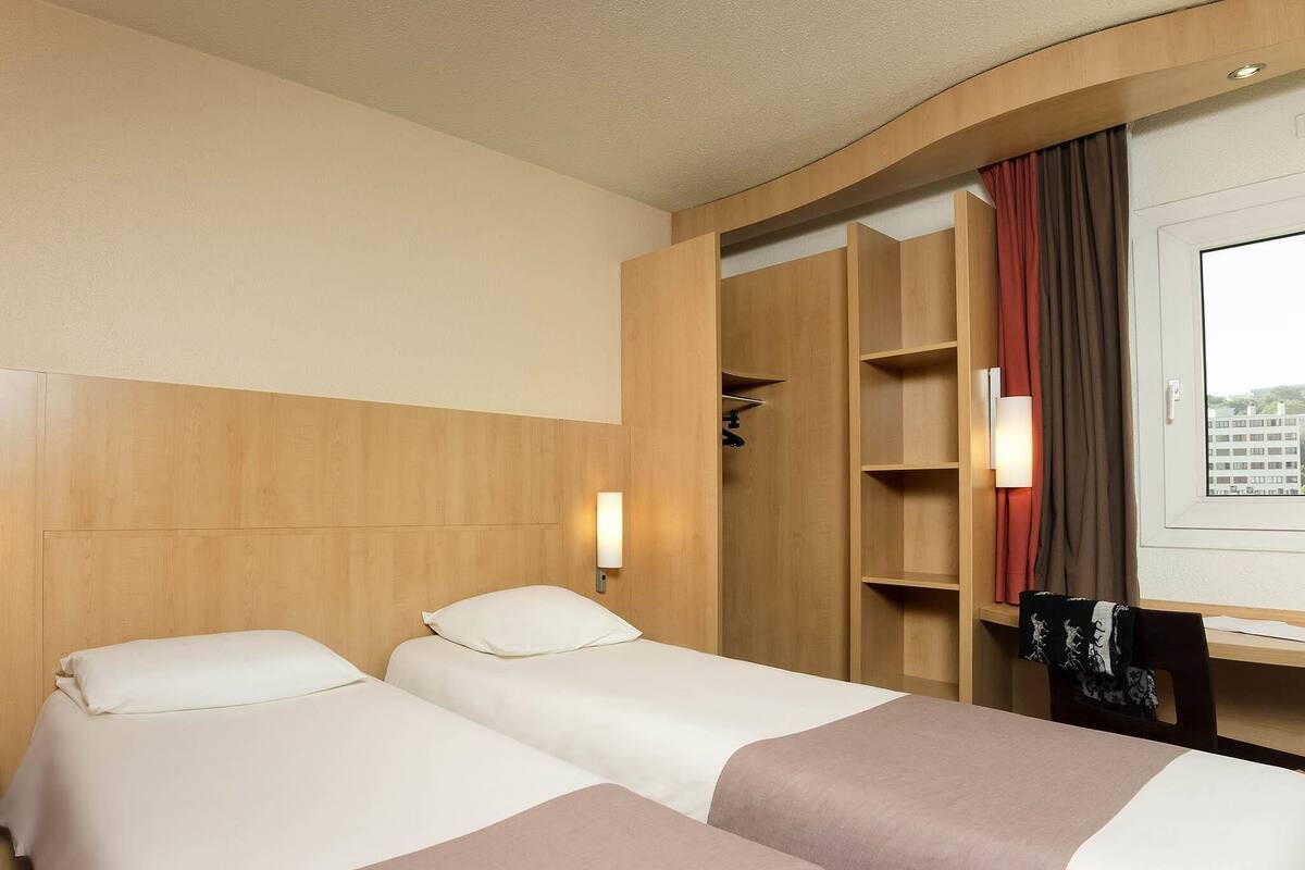 A hotel room with two beds, a wooden headboard, a desk with a chair, a window with curtains, and a wall-mounted light.