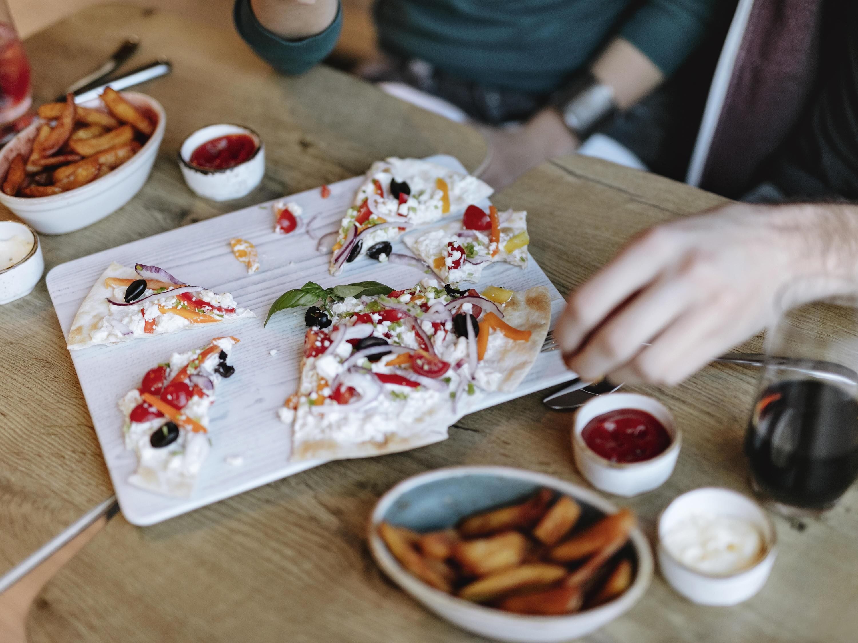 A dining table with a variety of food items including a pizza with toppings, a bowl of fries, and small bowls of sauces.