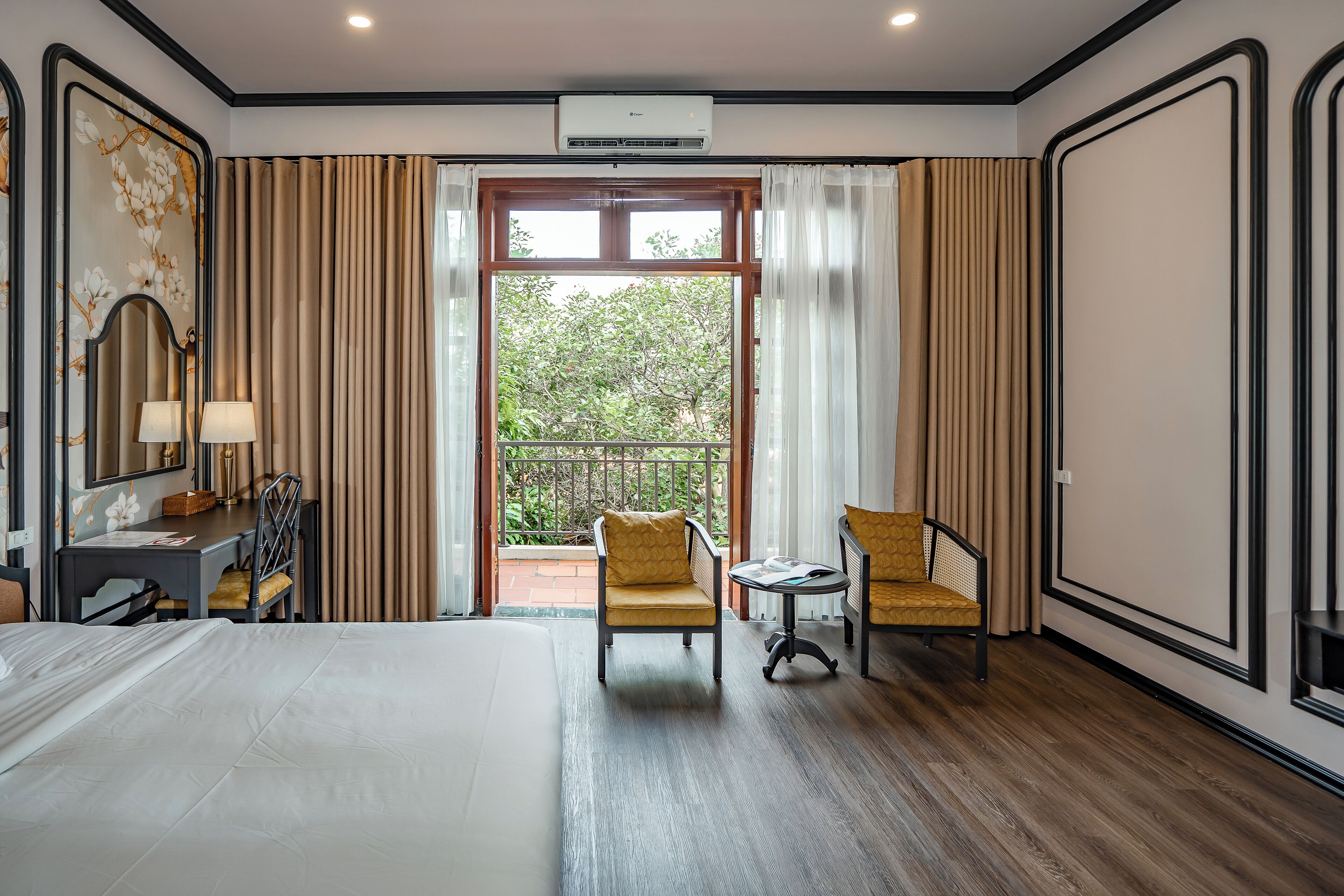 A hotel room with a large bed, a desk with a chair, two armchairs, a balcony with curtains, and an air conditioning unit.