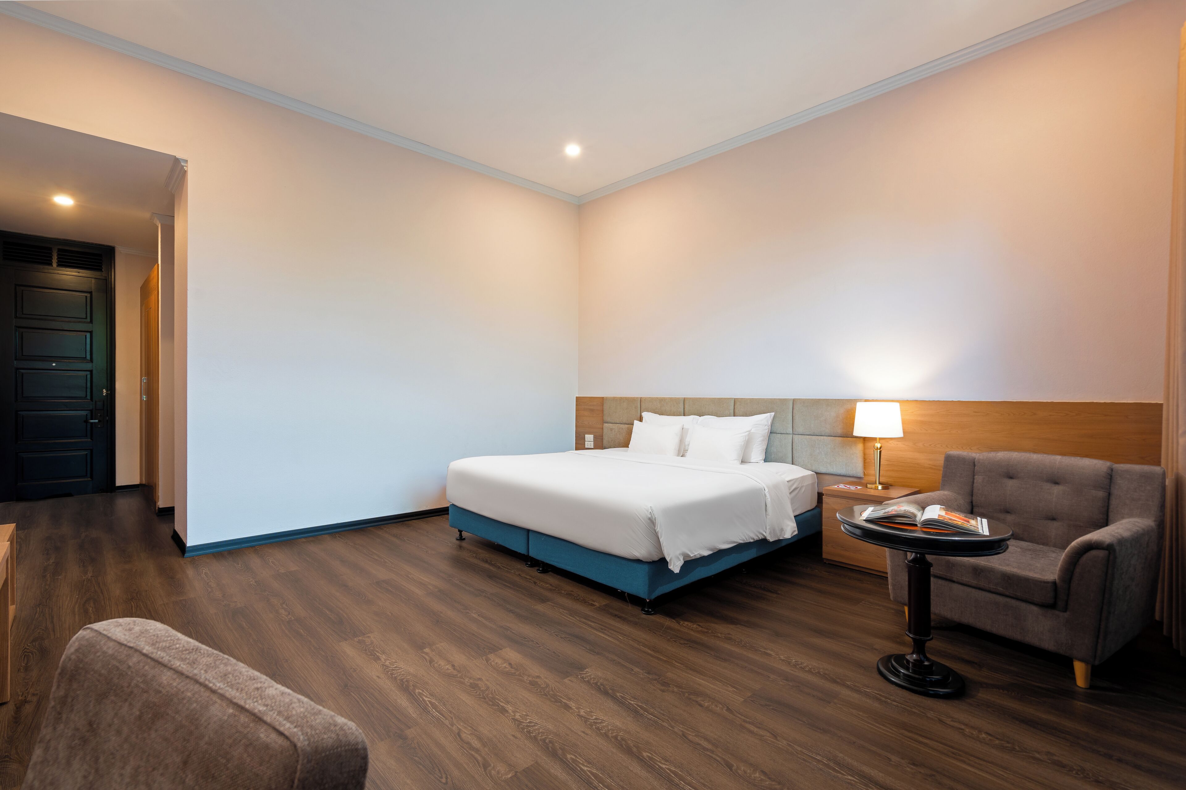 A hotel room with a bed, a chair, a small table with a lamp, and a wooden floor.