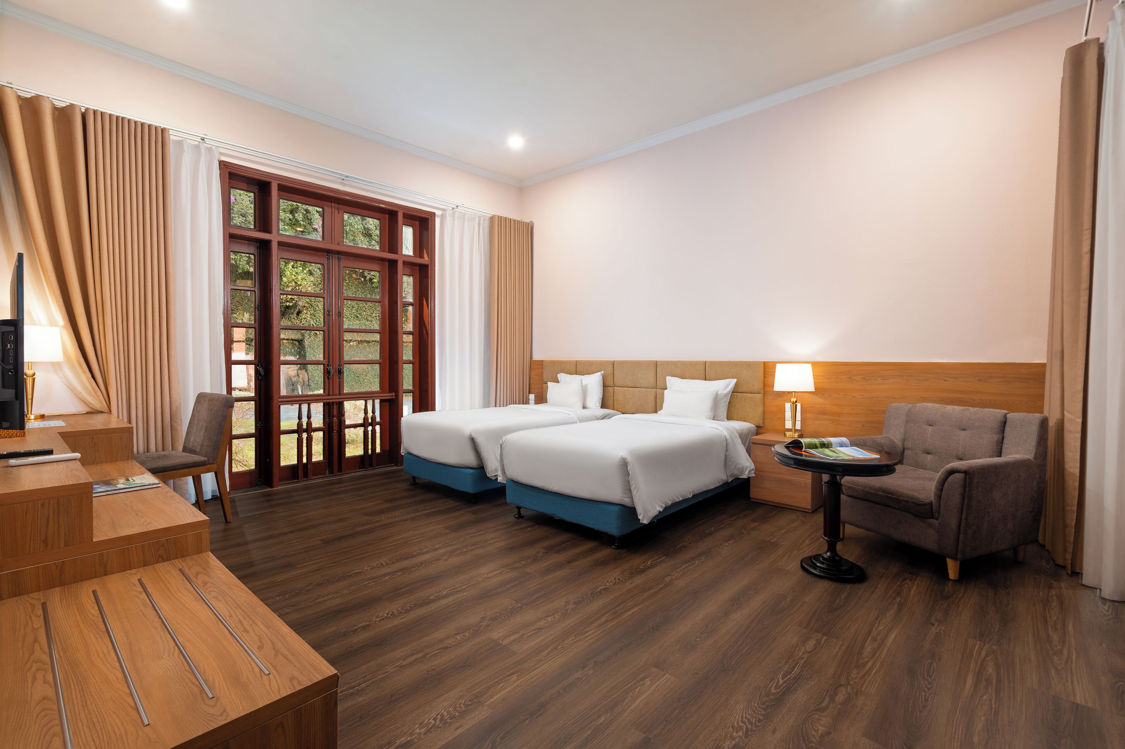 A hotel room with two beds, a desk, a chair, a table with a lamp, and a view of greenery through the window.