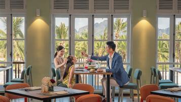 A dining area with tables and chairs, large windows with views of palm trees and mountains, and a person gesturing at a table.