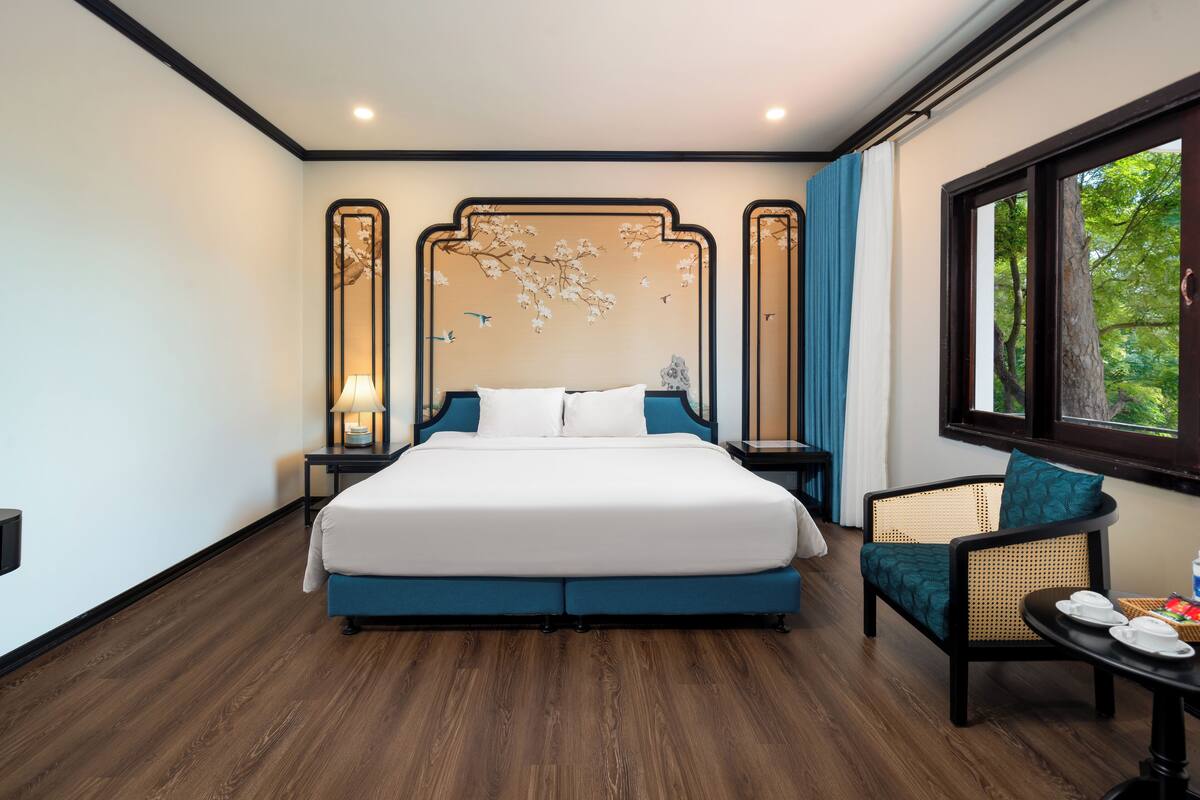 Una habitación de hotel moderna con una cama grande, una silla, una mesita de noche y una ventana con vistas a árboles.