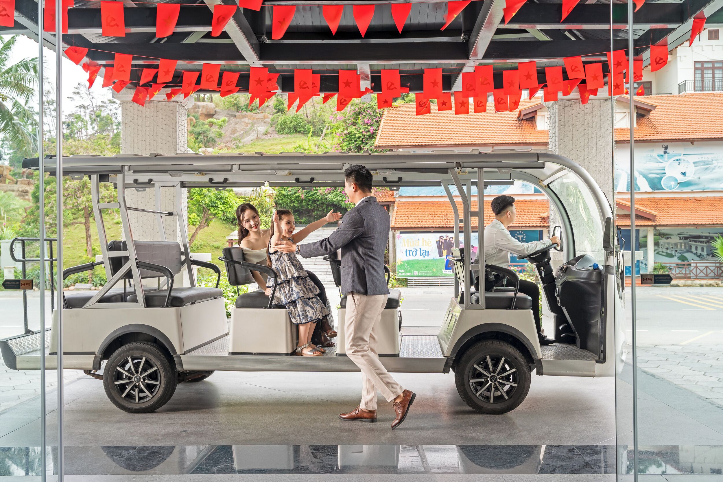 A golf cart with a driver and passengers, a glass barrier, and a building with a tiled roof in the background.