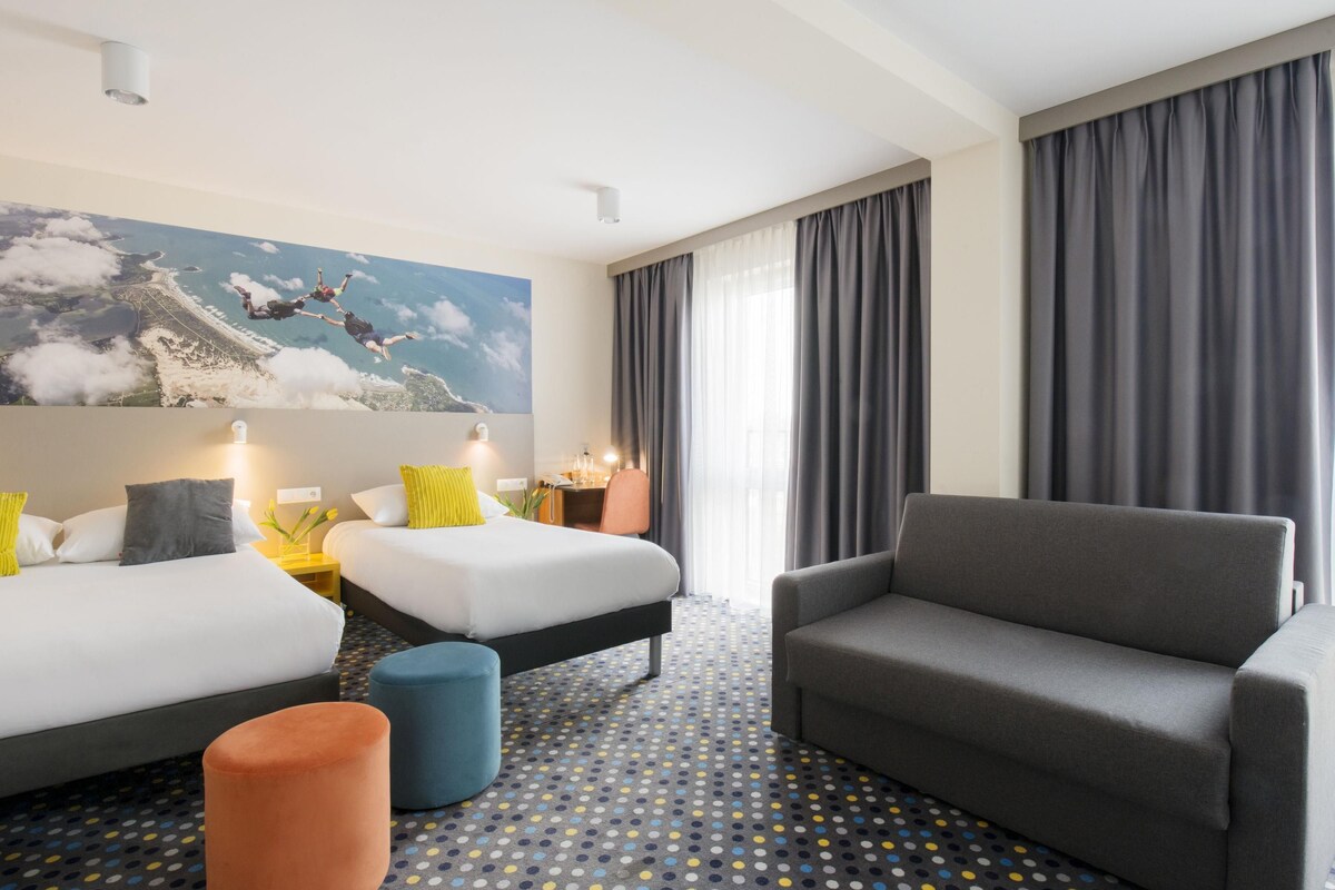 Habitación de hotel con dos camas, un sofá, un pequeño otoman, un escritorio y un ventanal con cortinas.