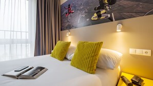 Una habitación de hotel con una cama grande, almohadas verdes, una revista, un teléfono y una mesita de noche con una lámpara y una billetera.