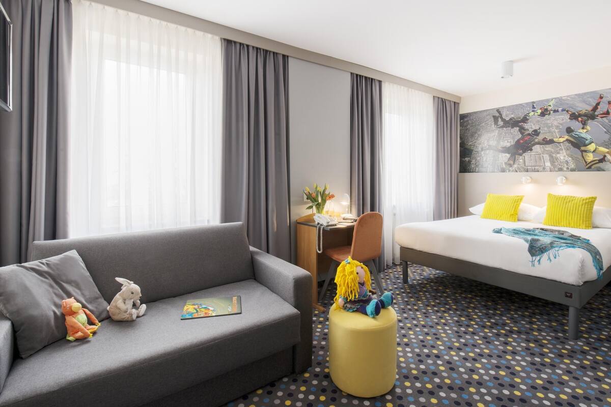 Een hotelkamer met een grijze bank, een klein rond voetenbankje, een bed met gele kussens, een bureau met lamp en een groot raam met grijze gordijnen.