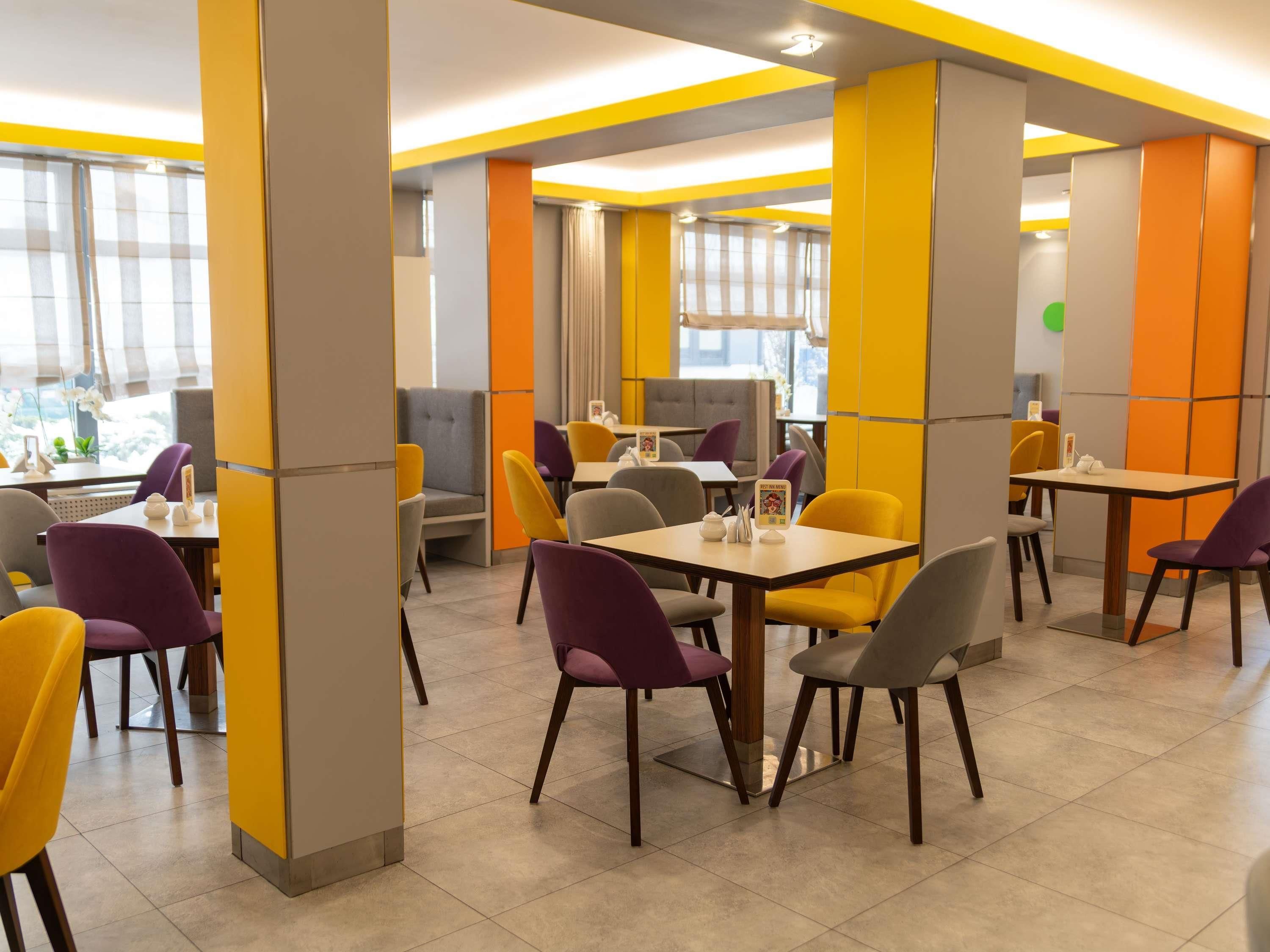 Un comedor moderno con paredes amarillas y grises, varias mesas y sillas, y vistas al exterior a través de amplios ventanales.