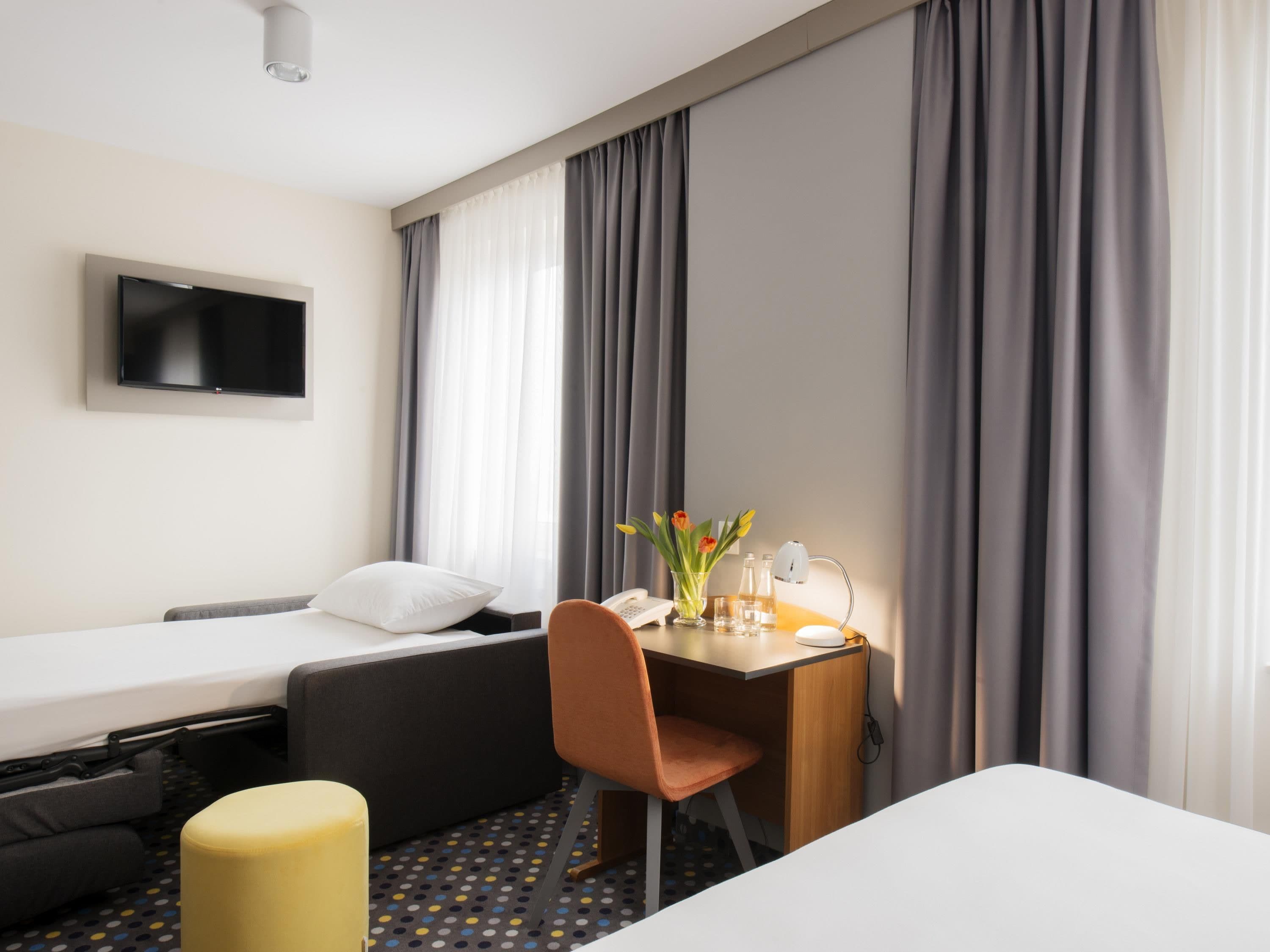 Ett hotellrum med en säng, ett skrivbord med en stol, en tv och ett fönster med gardiner.