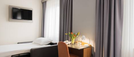 Ett hotellrum med en säng, ett skrivbord med en stol, en tv och ett fönster med gardiner.